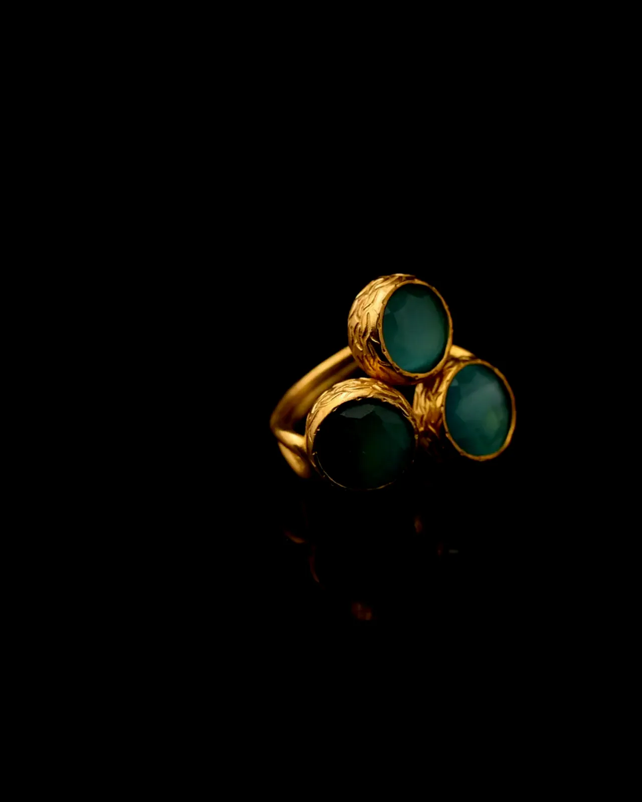 A gold-plated ring set with three teal stones, turning on black